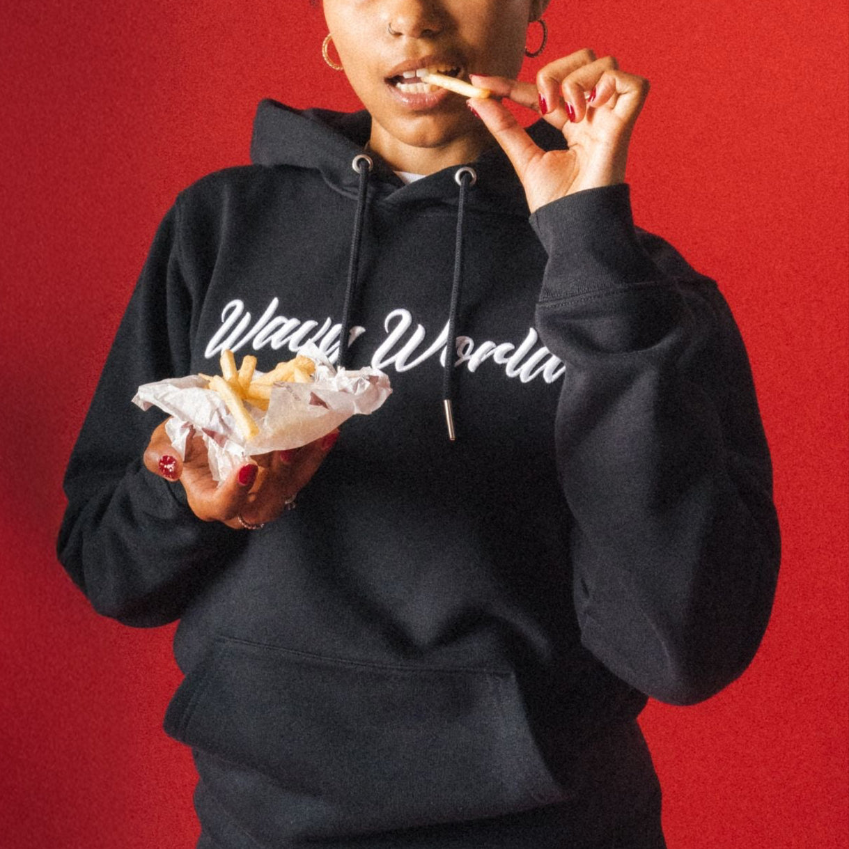 Person wearing the black Wavy World OG Hoodie from Drop 1 - Hip Hop & Cereal, featuring bold text, eating in front of a red background