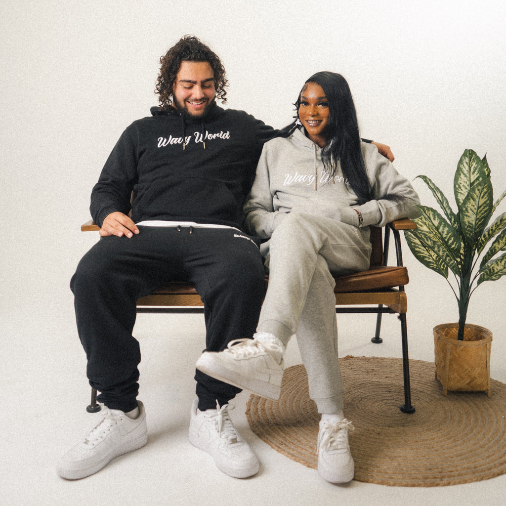 Two people wearing the full Wavy World OG Jogger set in black and grey, sitting side-by-side on a chair, showcasing relaxed streetwear style