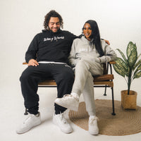 Two people wearing the full Wavy World OG Jogger set in black and grey, sitting side-by-side on a chair, showcasing relaxed streetwear style