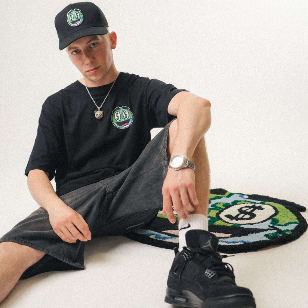 Person wearing a black Wavy World t-shirt, paired with shorts, sneakers, and a cap, sitting on the colourful Wavy World mascot rug.